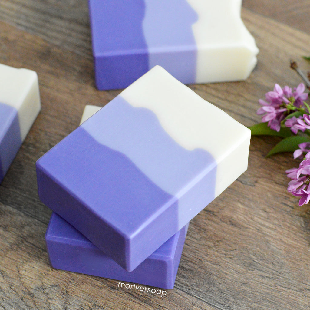 Image of Missouri River Soaps lilac soap in layers of purple, light purple, and white. Background is wood and real lilacs.