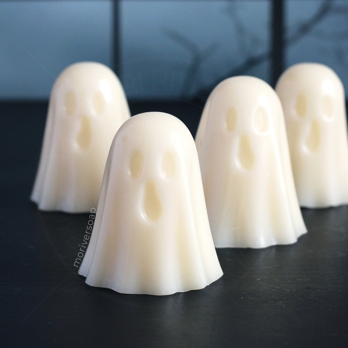 Four small white ghost-shaped wax melts with simple facial cutouts, arranged on a dark textured surface against a dark blurred background.