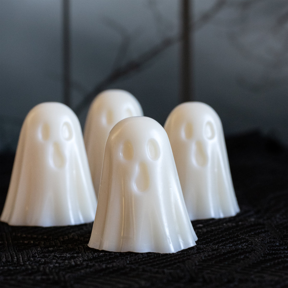 Four small white ghost-shaped wax melts with simple facial cutouts, arranged on a dark textured surface against a dark blurred background.