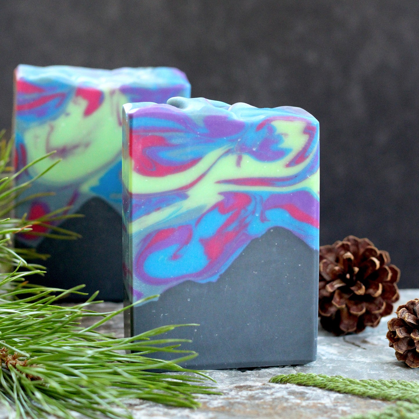 Missouri River Soap Northern Lights artisan soap with colorful swirls of blue, purple, pink, and green over a charcoal base, photographed with evergreen sprigs and pinecones on a rustic stone surface.