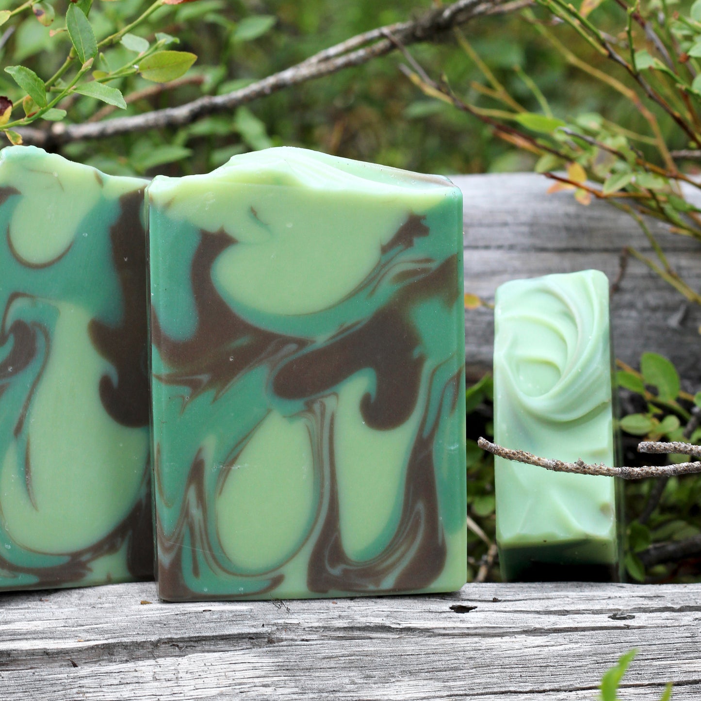 Missouri River Soap Territory: Bigfoot artisan soap with mint green and earthy brown swirls, photographed outdoors on a weathered log surrounded by forest greenery