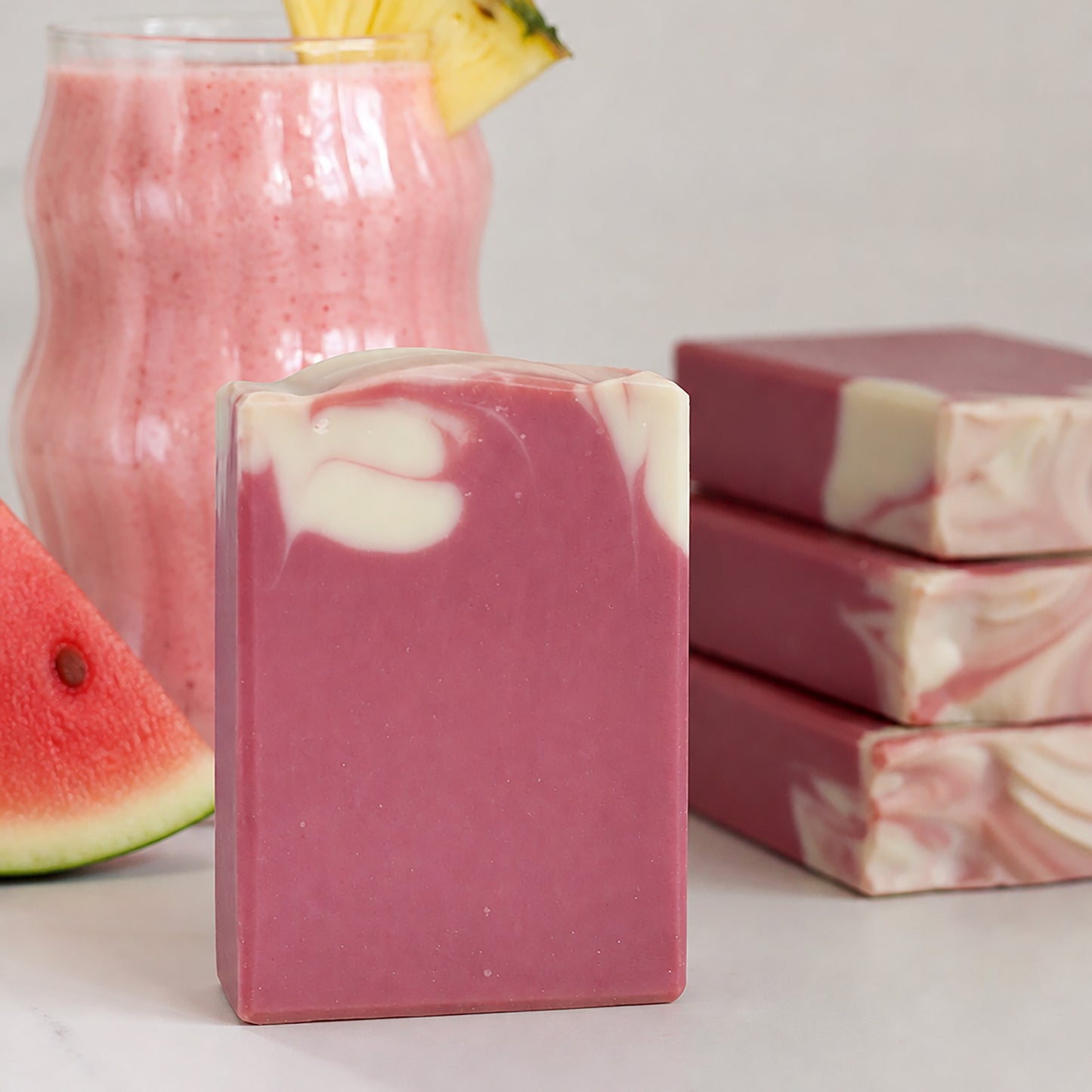Missouri River Soap Watermelon Paradise handcrafted cold process soap with pink and cream swirls, displayed beside a creamy watermelon, pineapple, and coconut drink with stacked soap bars in the background.