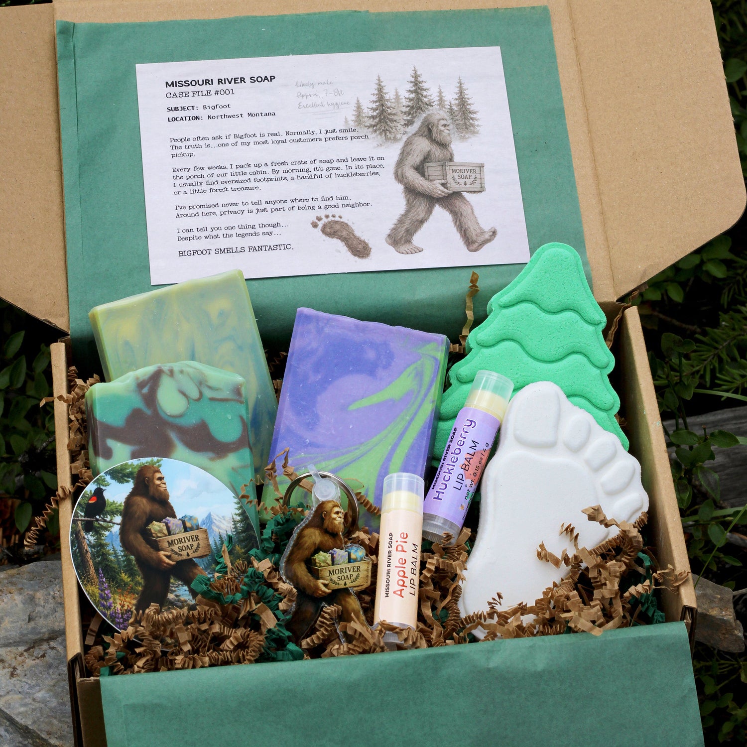 Missouri River Soap Bigfoot Bath & Body Box featuring three artisan soaps, two bath bombs, two lip balms, a Bigfoot acrylic keychain, sticker, and an exclusive Case File story card, arranged inside a kraft gift box with green tissue paper.