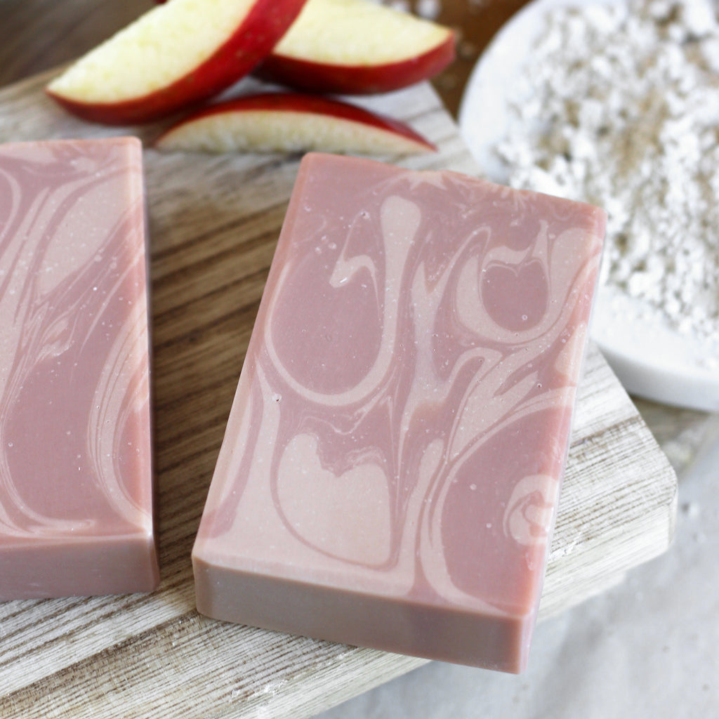 Two pink and tan soap bars on a wooden surface with apple slices in the background.