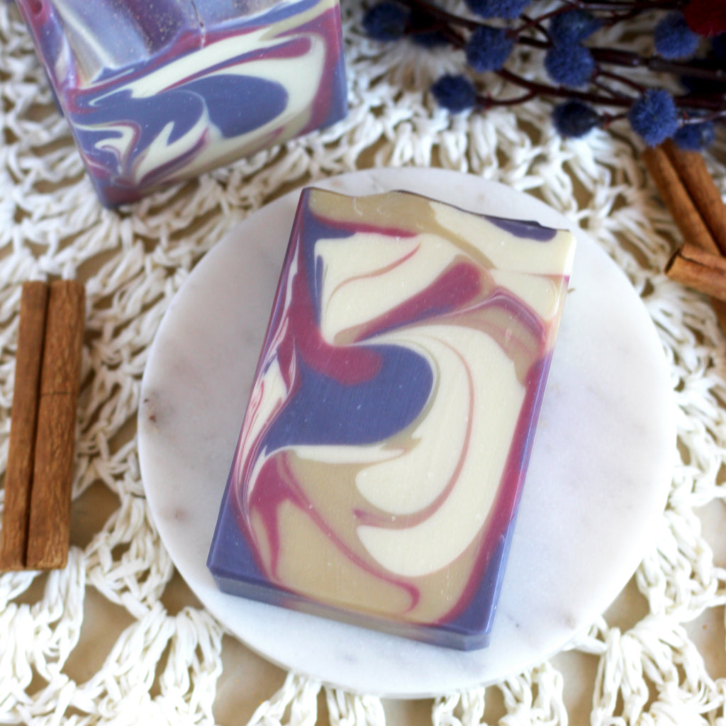 Soap bar with purple, pink, gold, and white swirls on a white plate, surrounded by cinnamon sticks.