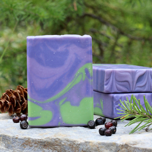 Missouri River Soap Bigfoot Bait artisan soap with purple and green swirls, photographed outdoors on a stone surface with huckleberries, evergreen sprigs, pinecones, and a forest backdrop
