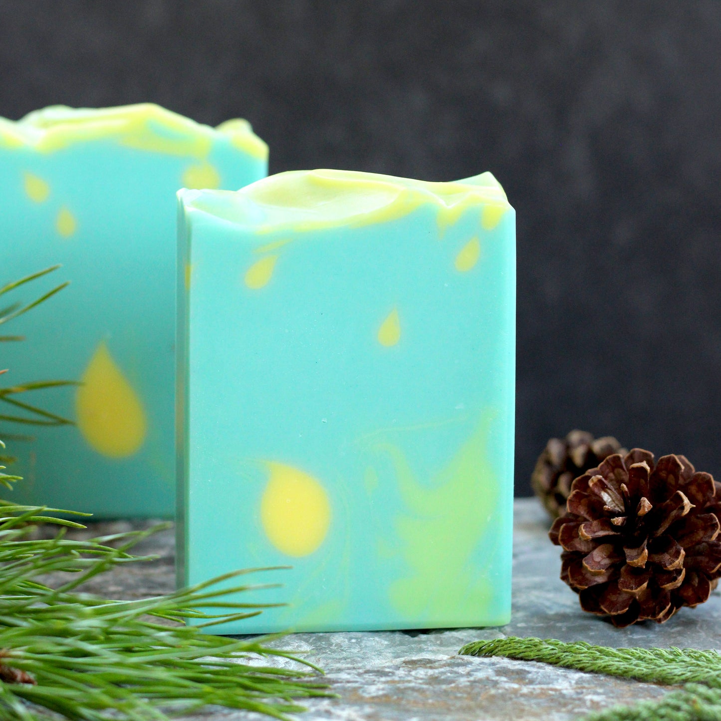 Missouri River Soap Chasing Fireflies artisan soap with a soft aqua design and glowing yellow accents, photographed with evergreen sprigs and pinecones on a rustic stone surfac