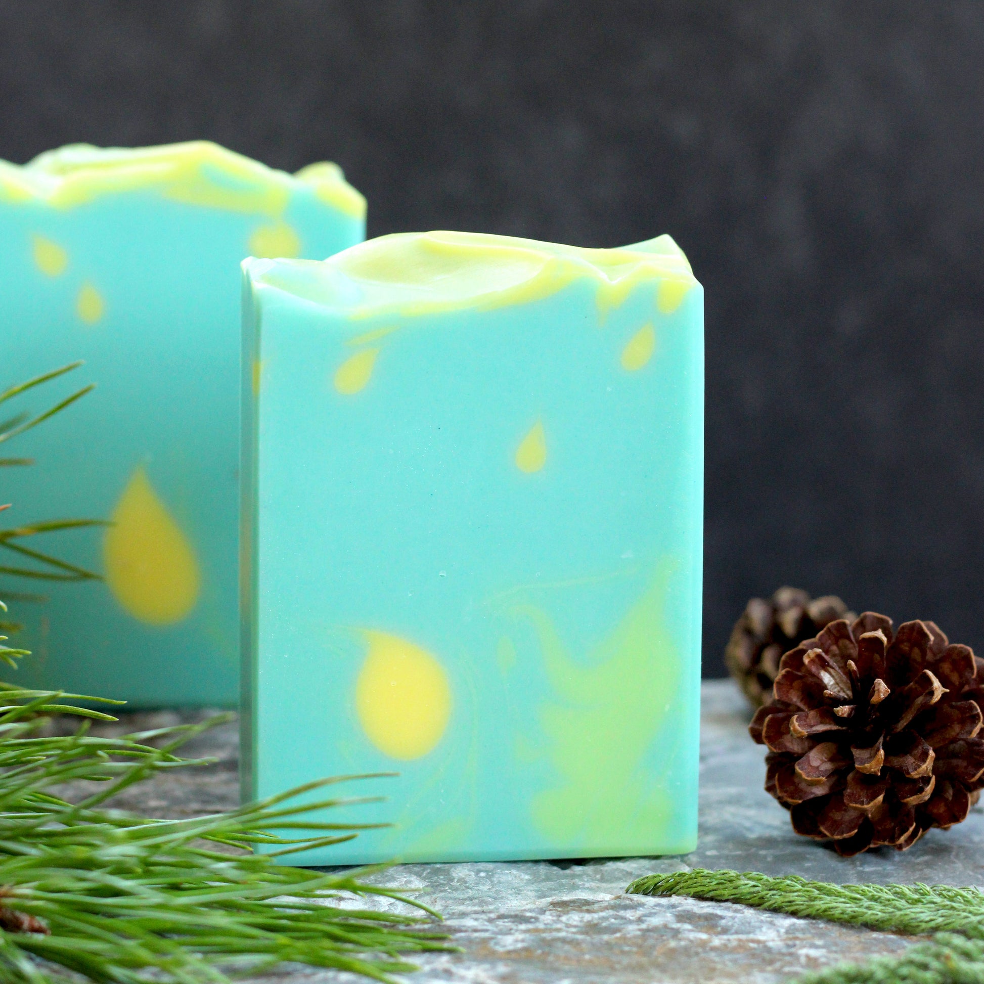 Missouri River Soap Chasing Fireflies artisan soap with a soft aqua design and glowing yellow accents, photographed with evergreen sprigs and pinecones on a rustic stone surfac