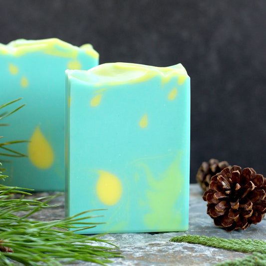 Missouri River Soap Chasing Fireflies artisan soap with a soft aqua design and glowing yellow accents, photographed with evergreen sprigs and pinecones on a rustic stone surfac