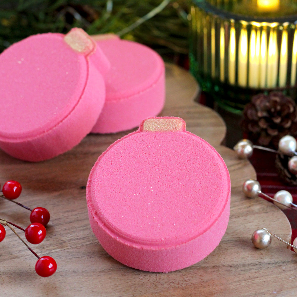 Missouri River Soap Cranberry & Balsam Bath Bomb – flat ornament-shaped bath bomb in reddish-pink with gold mica; festive fragrance of cranberry, plum blossom, mandarin, balsam, sage, and warm floral and woody notes.