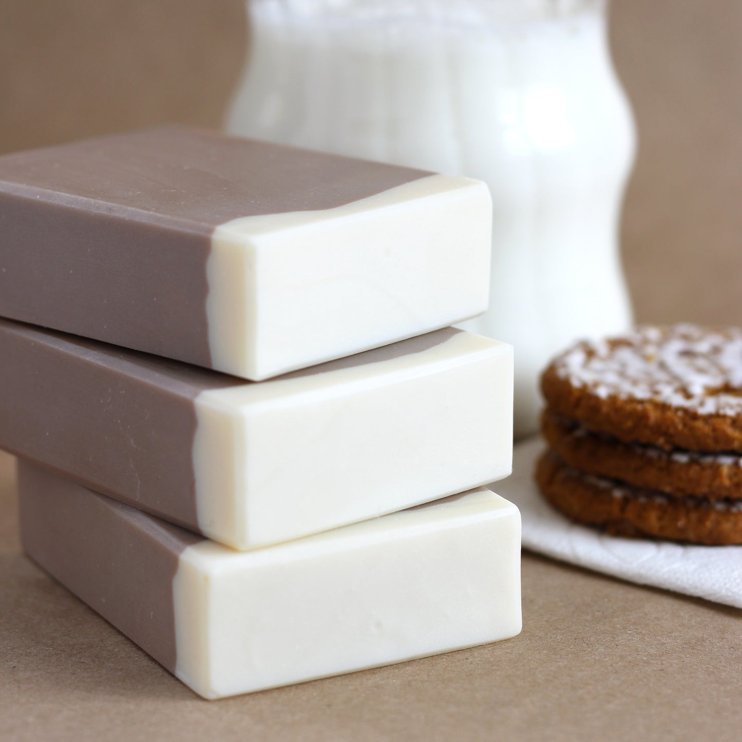 Three bars of soap stacked on a brown surface with milk and cookies in the background