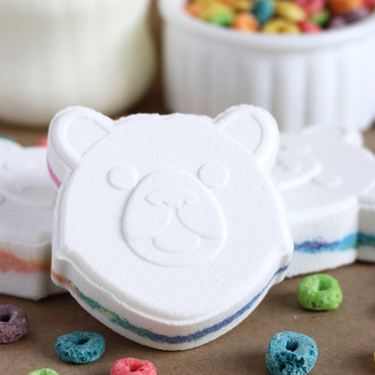 White bear-shaped bath bomb with rainbow center with colorful cereal pieces on a brown surface