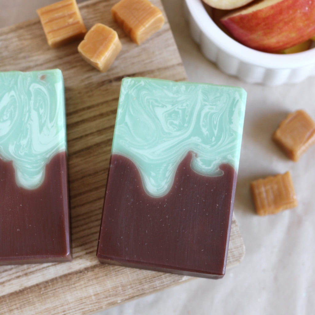 Two layered soap bars with a on a wooden board with caramel pieces and apple slices.