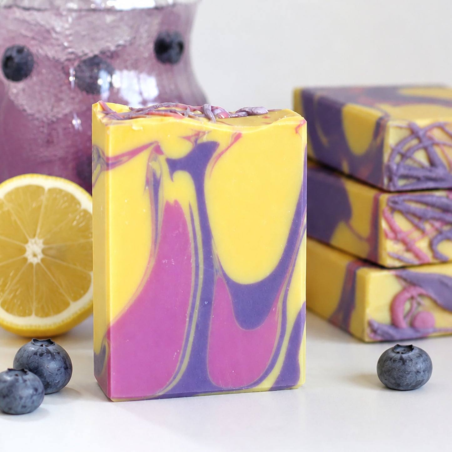 Handmade Berry Lemonade cold process soap with purple, pink, and yellow swirls displayed with blueberries, lemon, and a glass of berry lemonade by Missouri River Soap