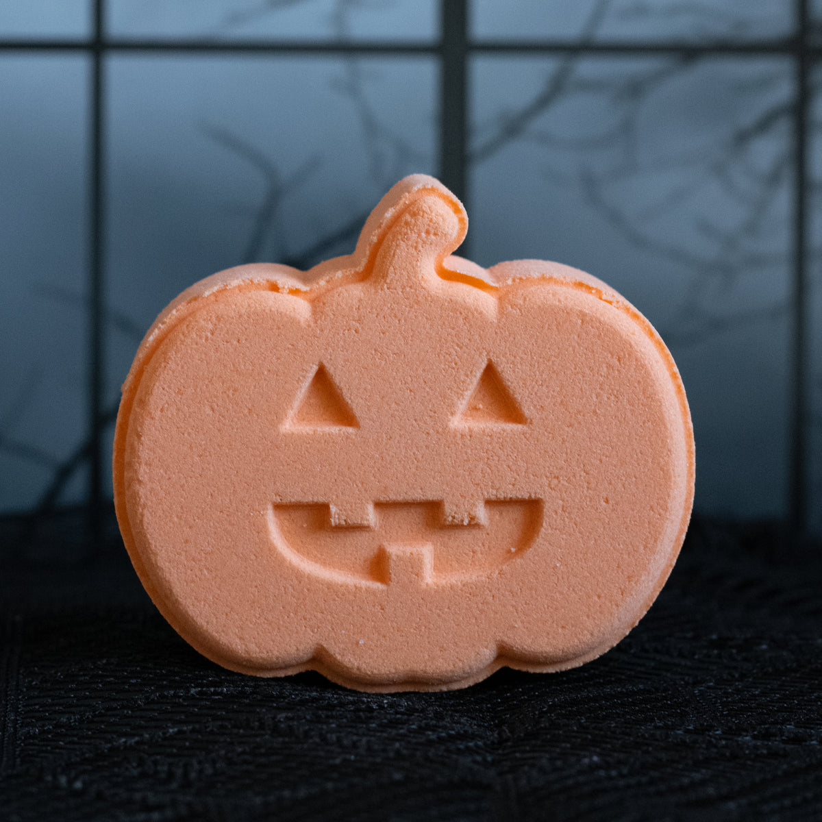 An orange pumpkin-shaped bath bomb with a carved jack-o'-lantern face, standing on a black textured surface against a dark, spooky background.