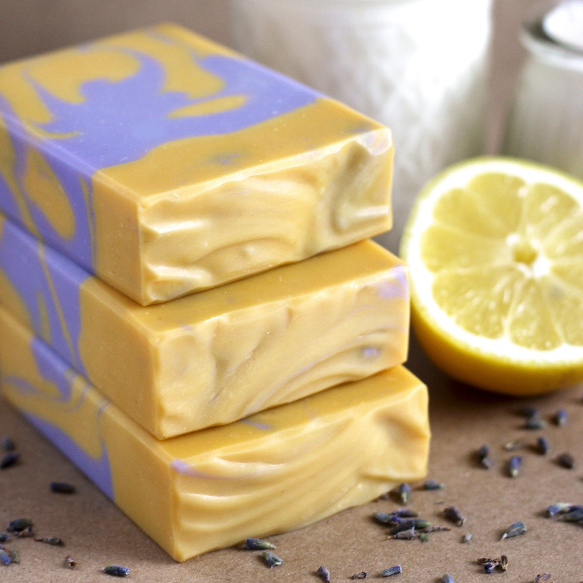 Three bars of yellow and purple swirled soap next to a lemon and lavender flowers on a brown surface.