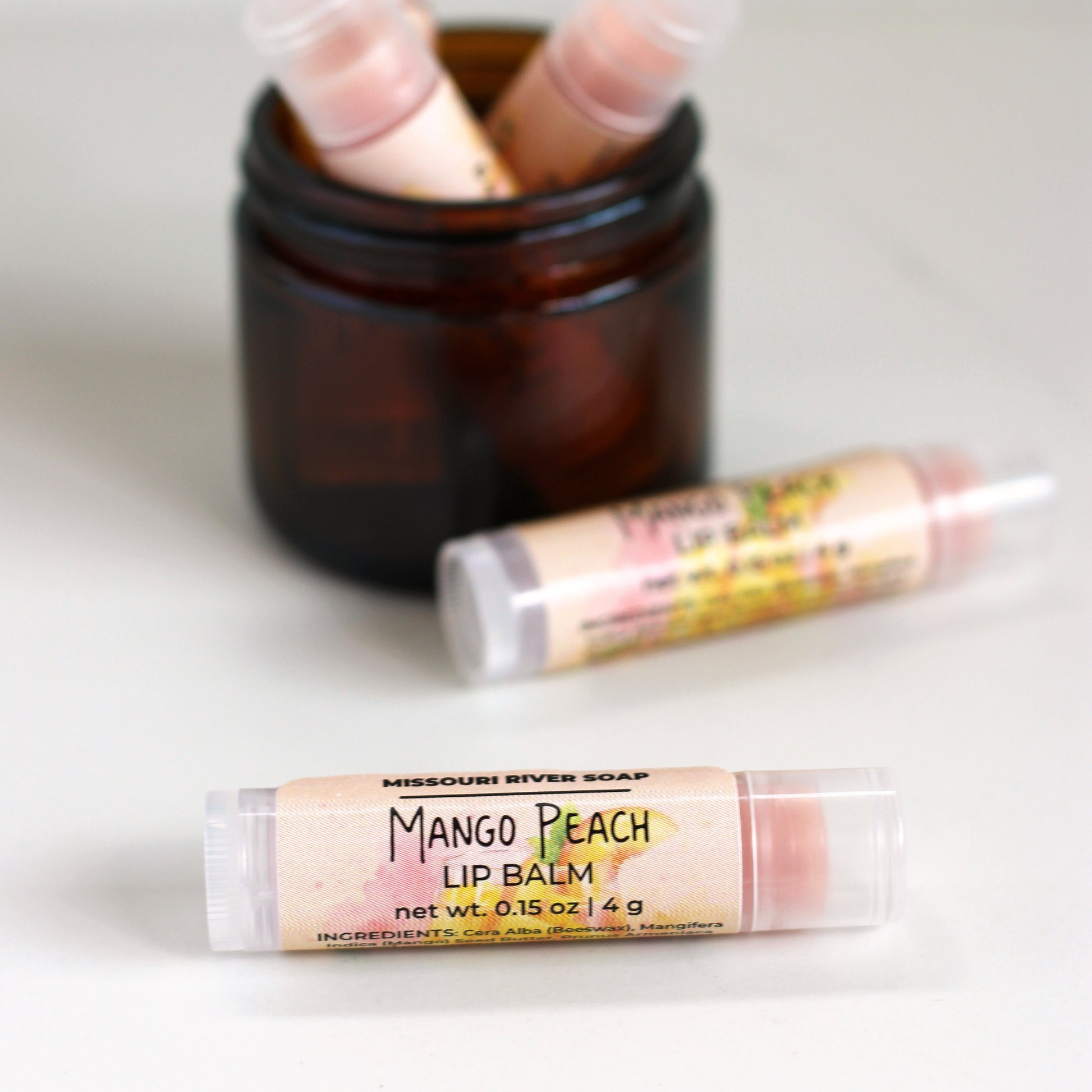 Mango Peach Lip Balm in a clear tube with a light peachy balm, handcrafted with mango butter, beeswax, and botanical oils. Features a bright mango peach fragrance with a subtle shimmer.