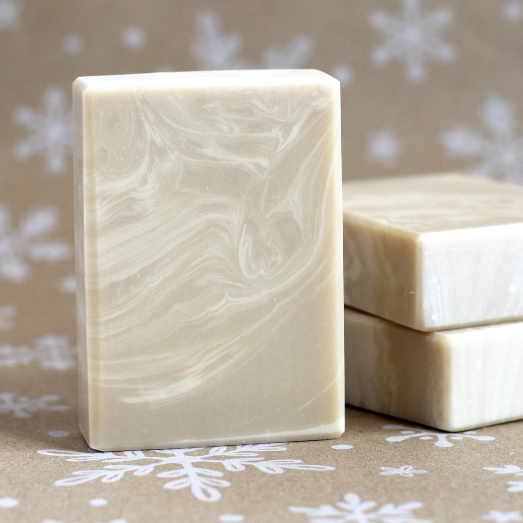 One bar of Milk & Honey soap in light tan with creamy swirls standing in front of two stacked bars, on a snowflake-printed kraft background