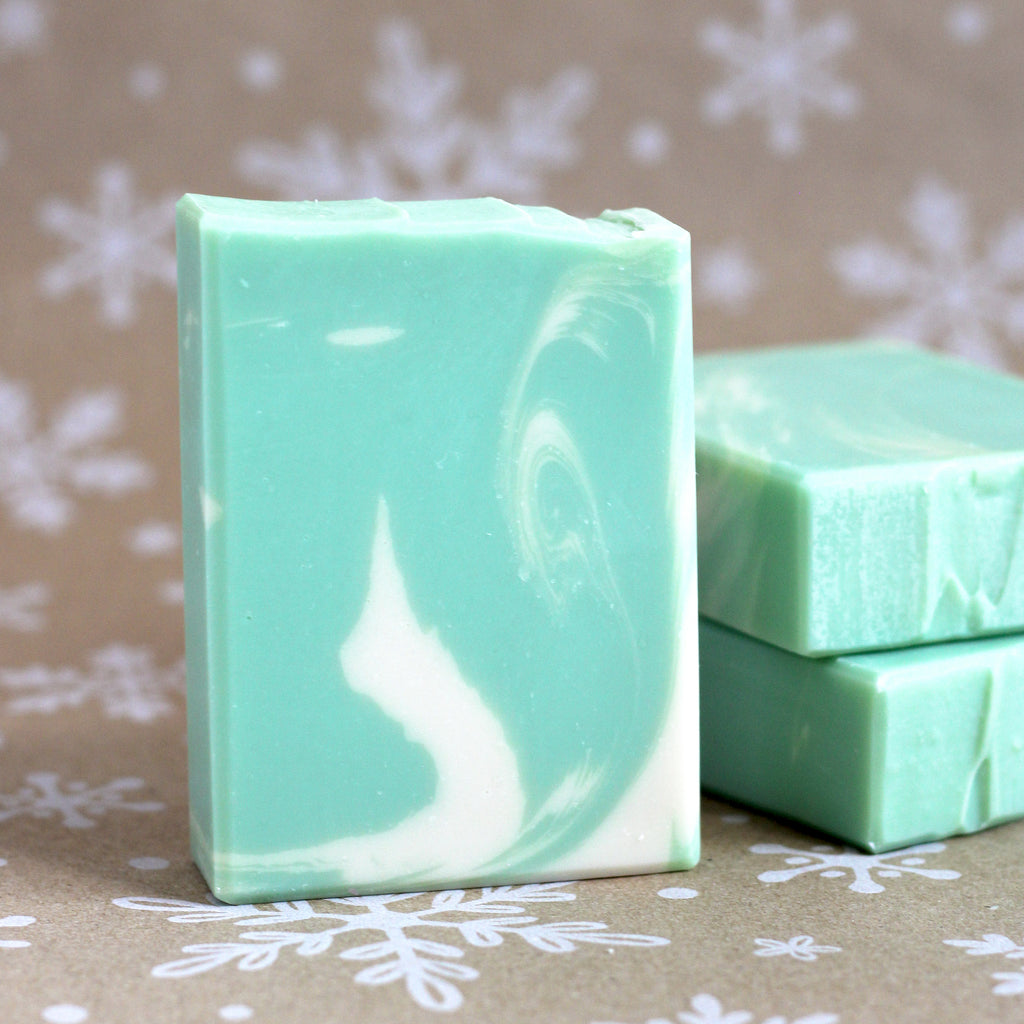 One bar of Minty Eucalyptus soap in mint green with creamy swirls standing in front of two stacked bars, on a snowflake-printed kraft background
