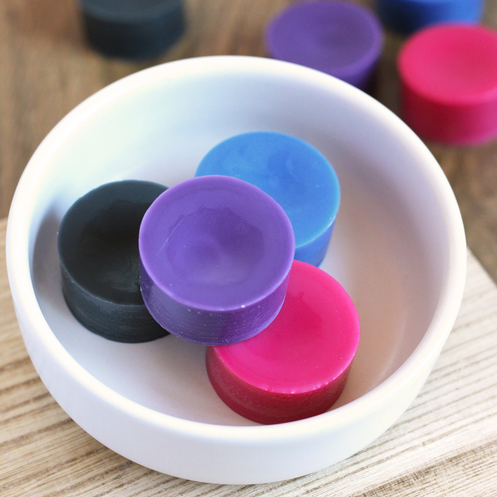 Purple, pink, blue, and black wax melt disks in a bowl on a wooden surface