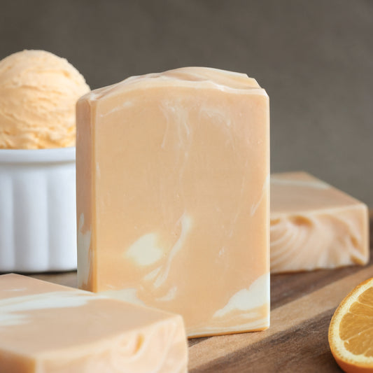 A rectangular bar of marbled soap in a warm peachy beige color stands on a wooden surface, with additional soap bars and a scoop of orange ice cream in a white bowl blurred in the background. A slice of orange is visible at the lower right.