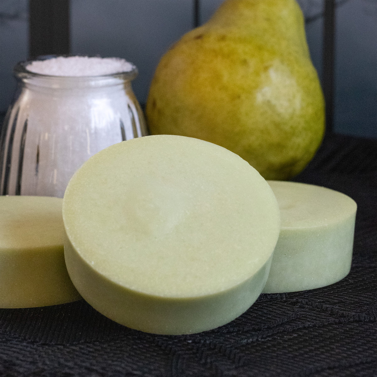 Pear-scented sea salt soap bars with a fresh pear and jar of sea salt against a moody window backdrop