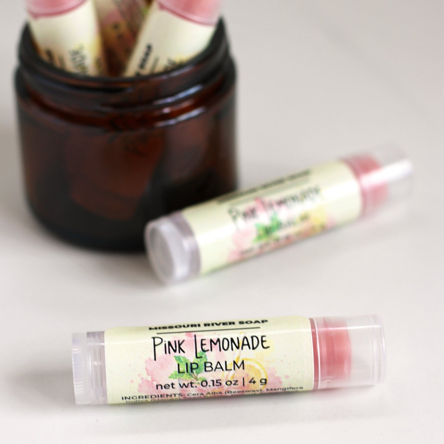 Pink Lemonade Lip Balm in a clear tube with a light pink balm, handcrafted with mango butter, beeswax, and botanical oils. Features a bright berry and pink lemonade fragrance with a subtle shimmer.