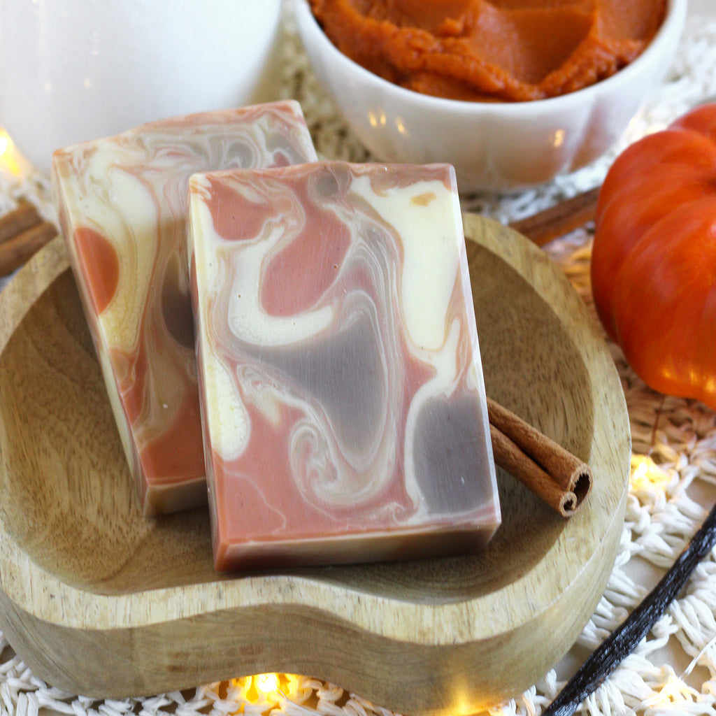 Two swirled soap bars on a wooden plate with pumpkins and cinnamon sticks in the background.