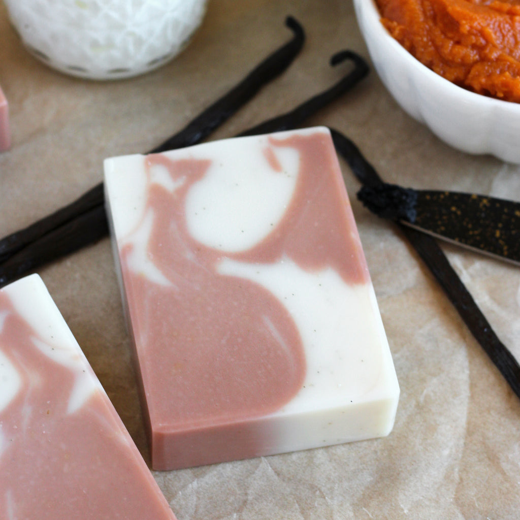 Two bars of orange and white soap with vanilla beans and a bowl of pumpkin on a beige surface.