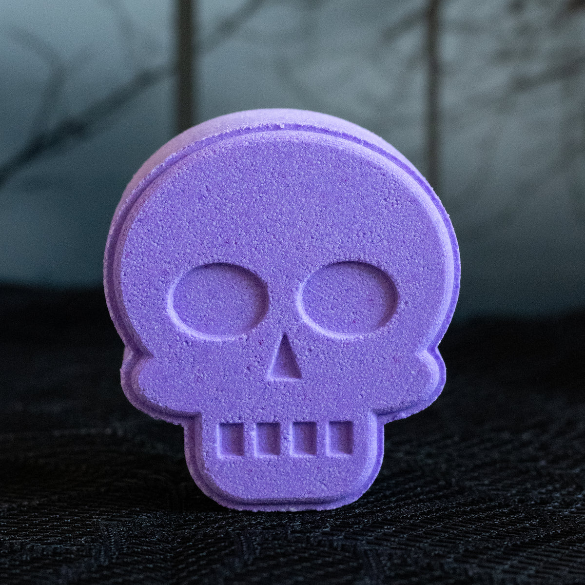 A purple skull-shaped bath bomb standing upright on a dark textured surface with a blurred dark background.