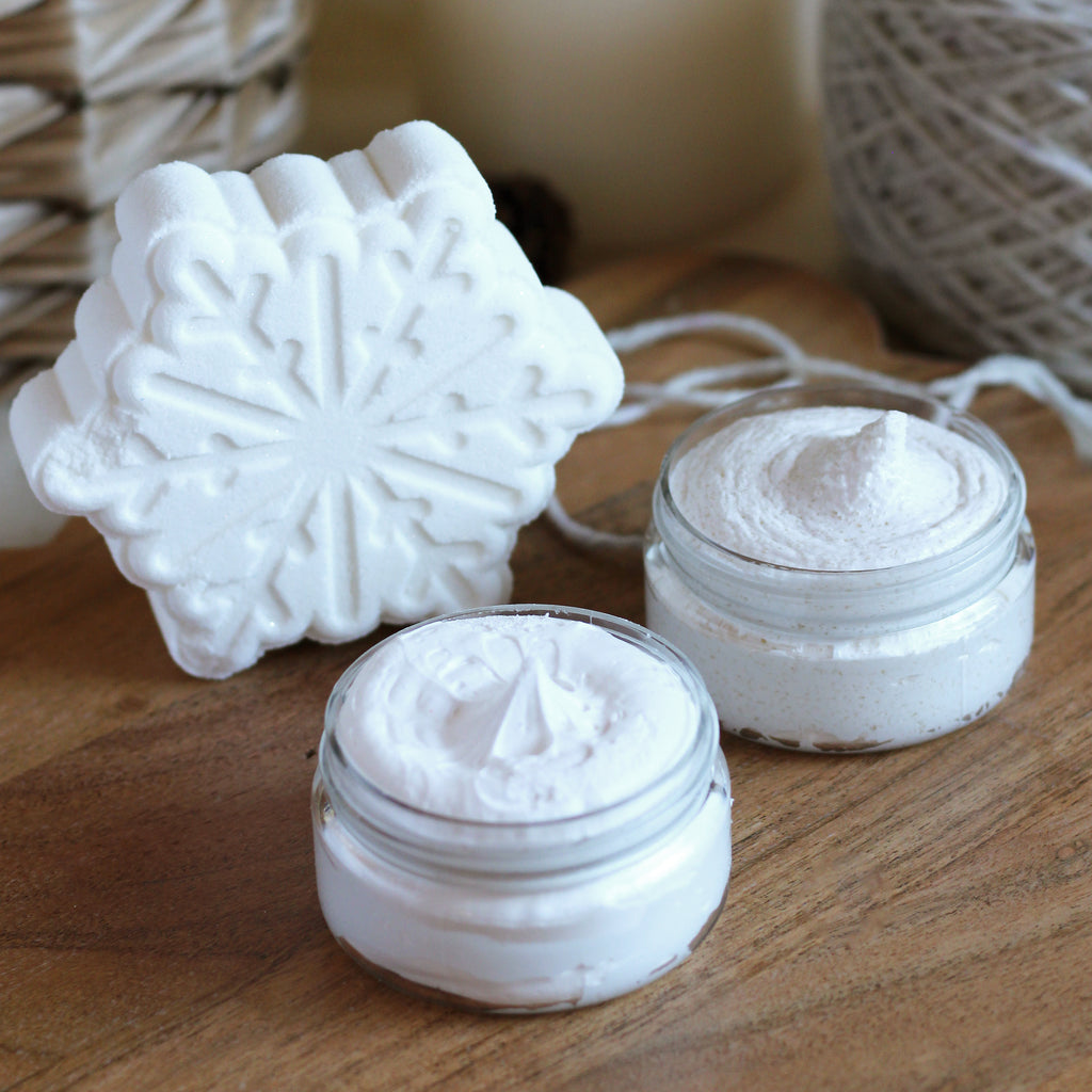 White snowflake bath bomb and two mini jars with cream soap and sugar scrub on a wood surface.