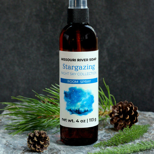 Missouri River Soap Stargazing Room Spray in an amber bottle with a blue night sky label, photographed on a stone surface with evergreen sprigs and pinecones