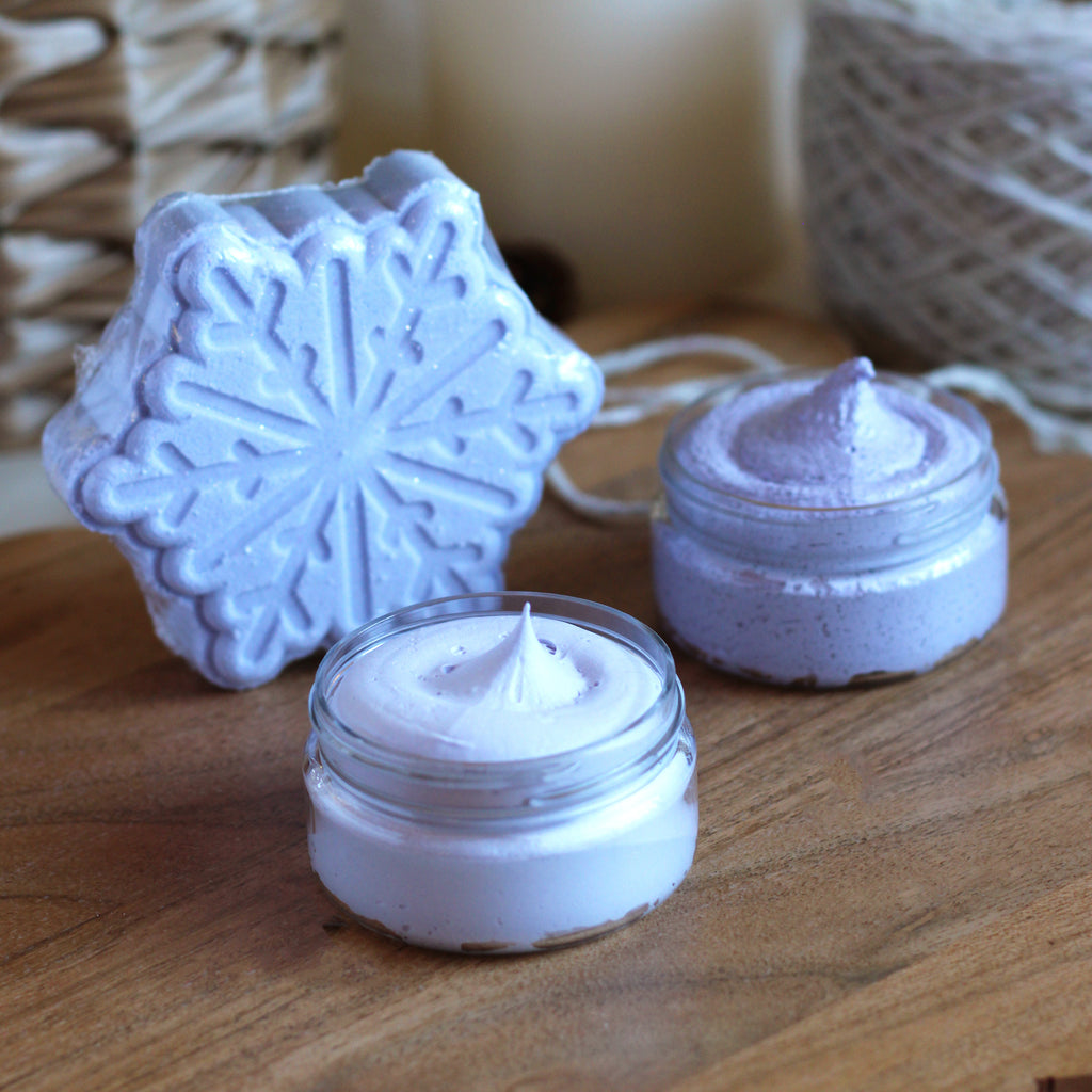 Bluish-purple snowflake bath bomb and two mini jars with cream soap and sugar scrub on a wood surface.