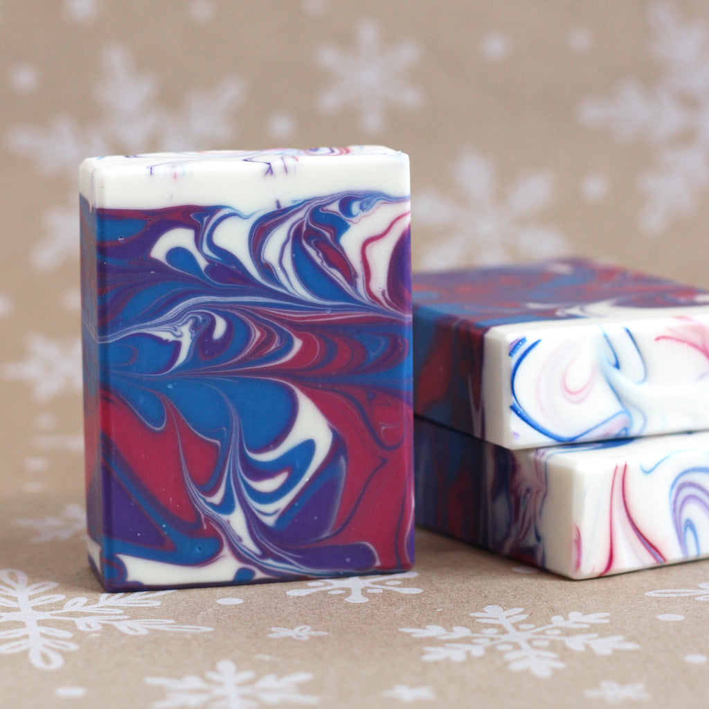 One bar of Wild Berry Blizzard soap with swirls of pink, blue, purple, and white standing in front of two stacked bars, on a snowflake-printed kraft background