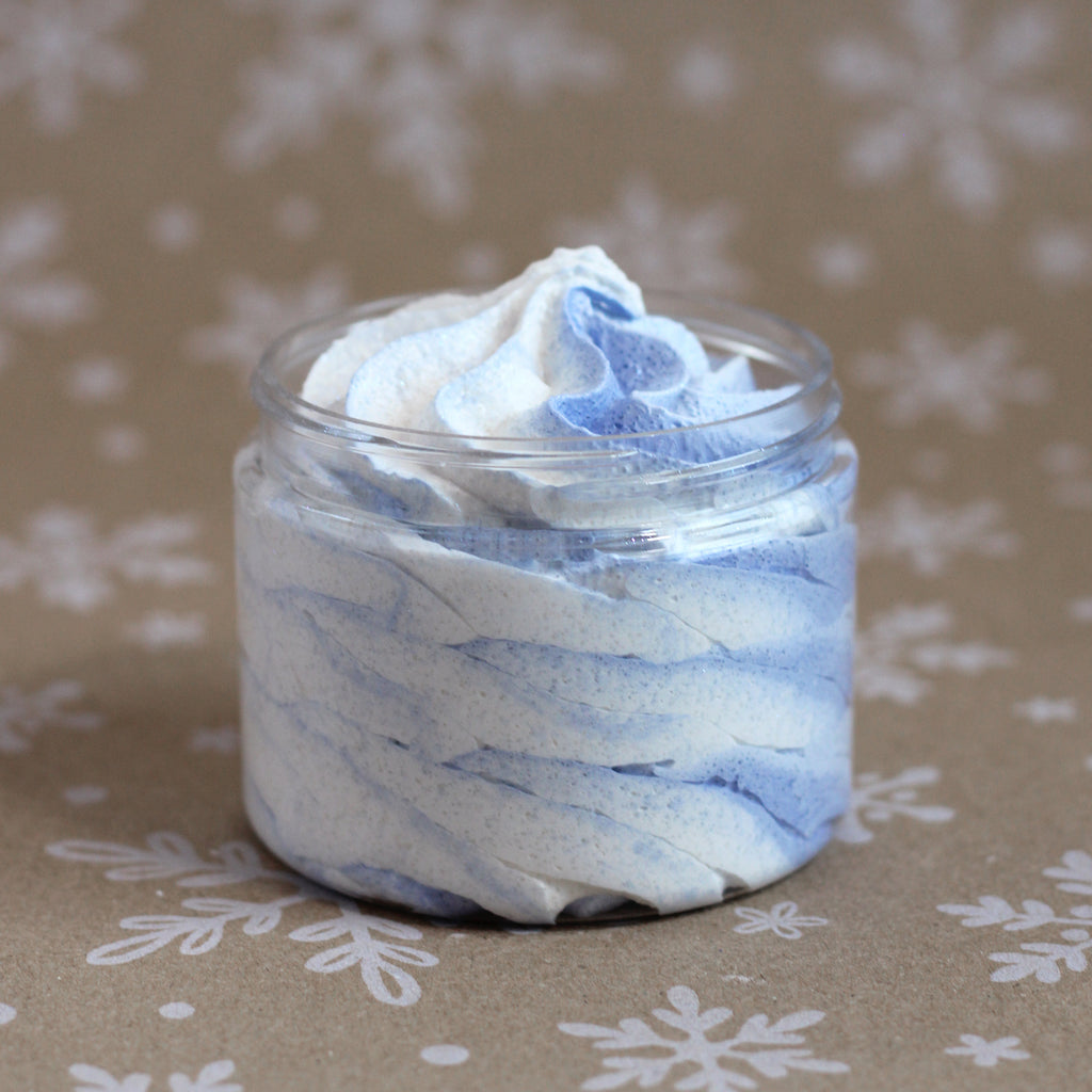 Winter Wonderland whipped soap sugar scrub in a clear jar with blue and white swirls, set on a kraft snowflake background.