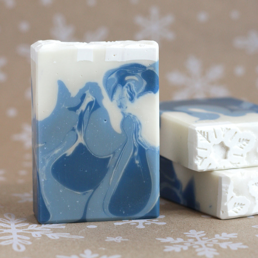 One bar of Winter Wonderland soap with swirls of dusky blue and creamy white, embedded with white snowflakes, standing in front of two stacked bars, on a snowflake-printed kraft background